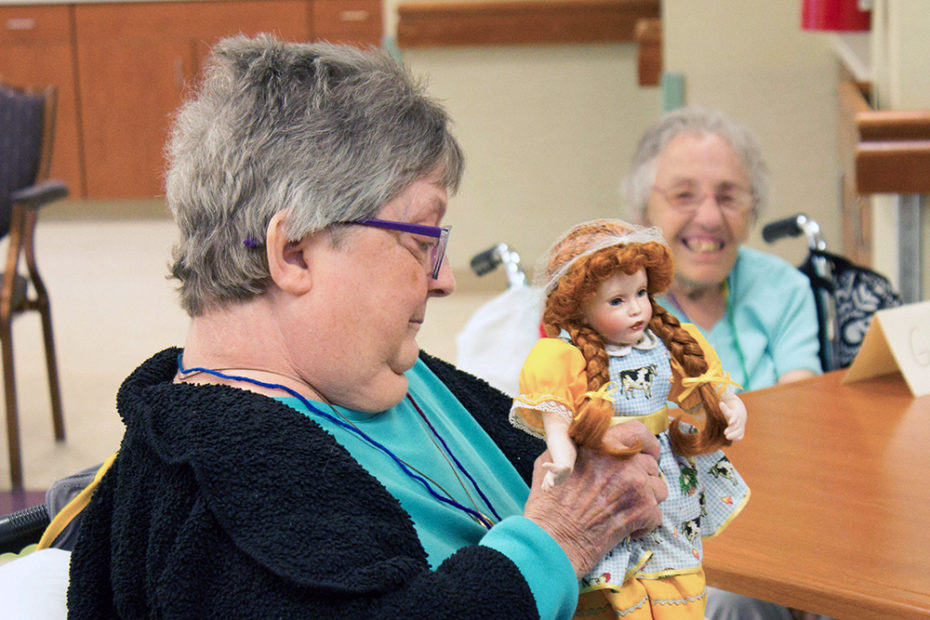 Two participants smiling as they look at one participant's doll with red braids with a yellow dress and a blue and white checkered apron with a cow on the front.